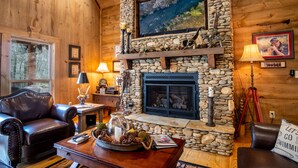 A cozy log cabin interior with a stone fireplace, leather furniture, and a scenic view through the window.