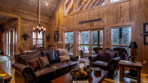 A spacious living room with leather sofas, a coffee table with a decorative bowl, and a chandelier. Large windows provide natural light and views of the outdoors.