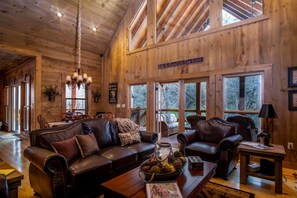 A spacious living room with leather sofas, a coffee table with a decorative bowl, and a chandelier. Large windows provide natural light and views of the outdoors. - Amazing Luxury Getaway ~ 50 Feet on the Toccoa River ~ Great Fishing (BLUE RIDGE)