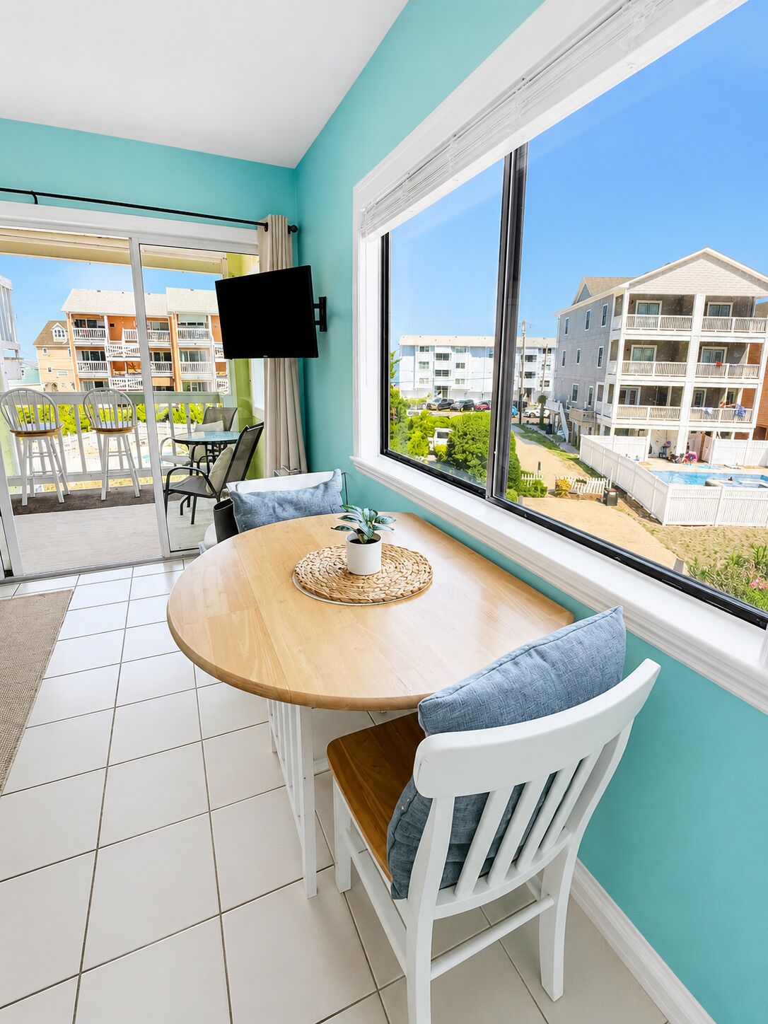 A room with a round wooden table, white chairs, a TV mounted on the wall, and a large window with a view of buildings and a swimming pool.