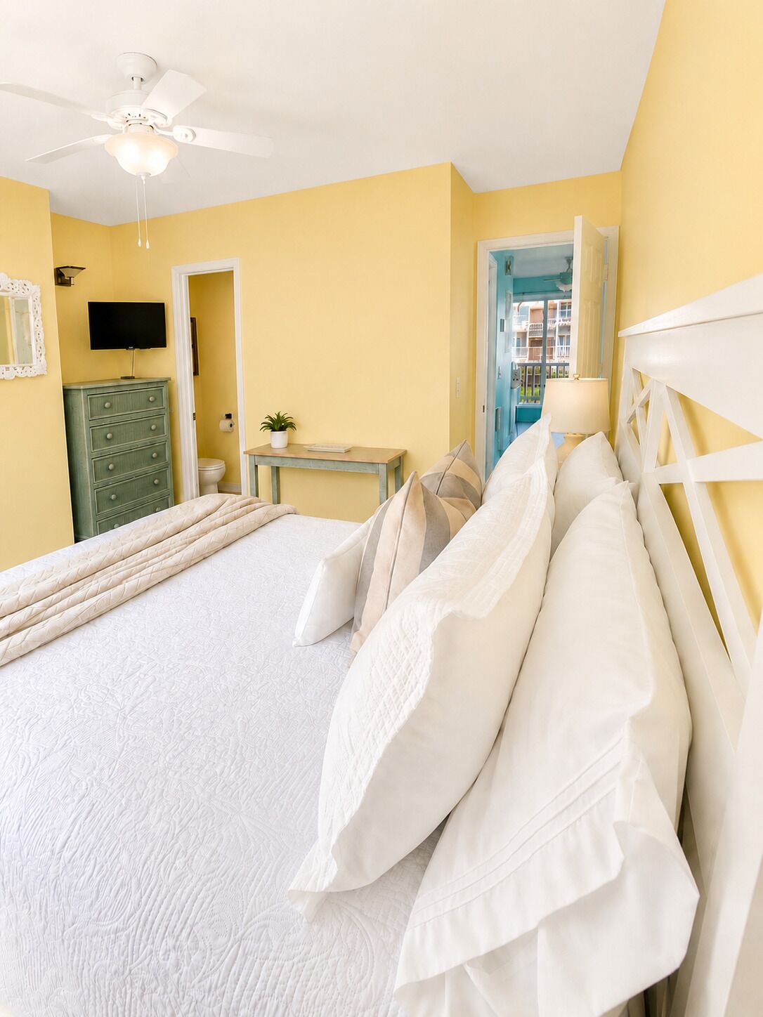 A bedroom with a bed, a ceiling fan, a TV, a dresser, and a small table with a plant.
