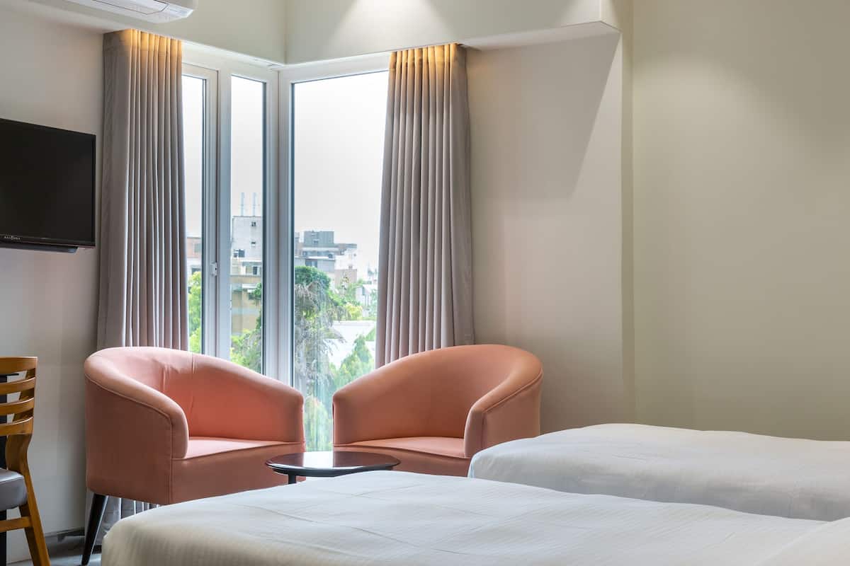 A hotel room with two beds, a ceiling fan, a TV mounted on the wall, and a view of the city through large windows.