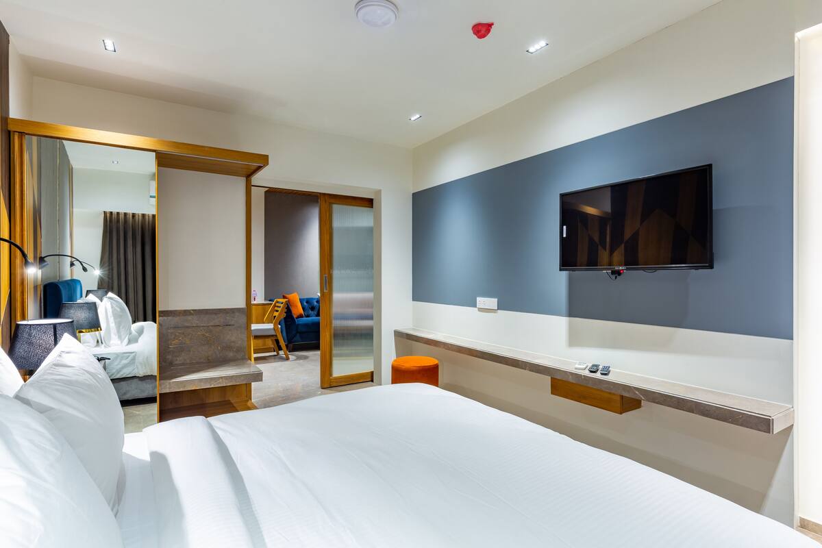 A modern hotel room with a large bed, a TV mounted on the wall, a small table, and a seating area with a sofa and chairs.