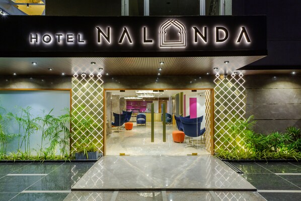 La entrada del Hotel Nalonda, con un diseño moderno, presenta un letrero destacado, una puerta de vidrio y un área de descanso con sillas de colores.