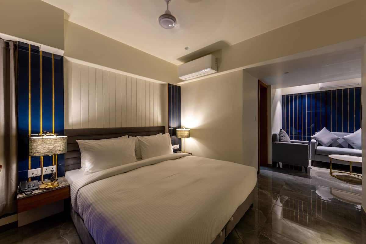A modern hotel room with a large bed, bedside lamps, a nightstand, a desk, and a seating area with a sofa and armchairs.