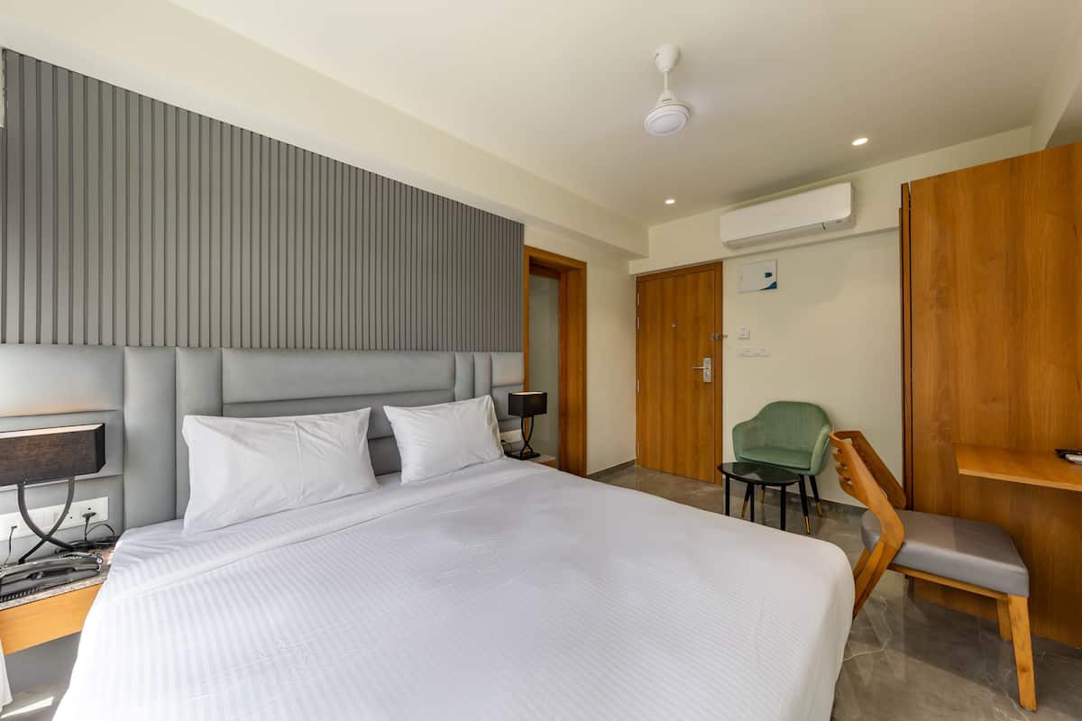 A modern hotel room with a large bed, a desk, a chair, a small table, and a wall-mounted air conditioner.