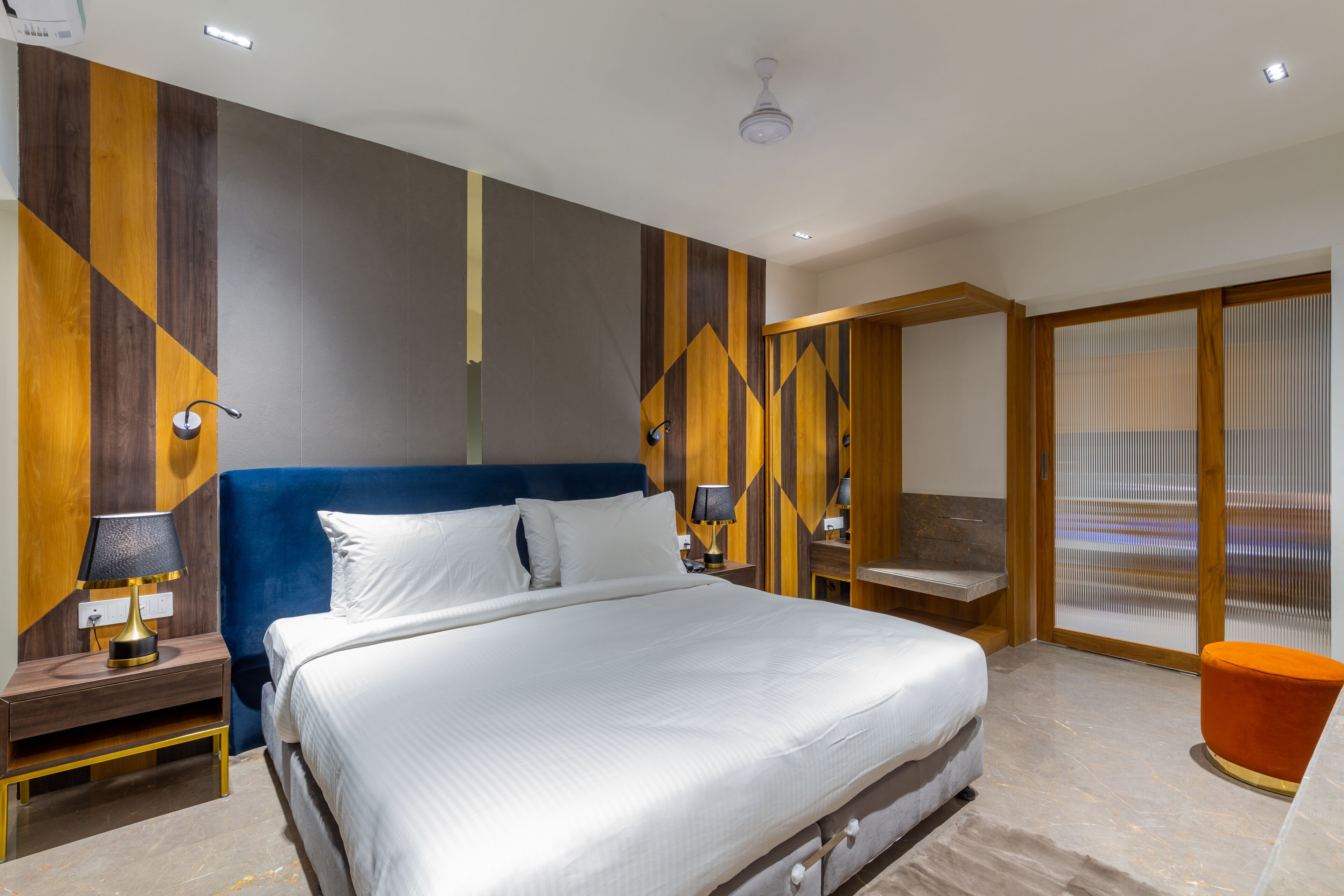 A modern hotel room with a large bed, a nightstand with a lamp, a wooden headboard, and a view of the city through large windows.