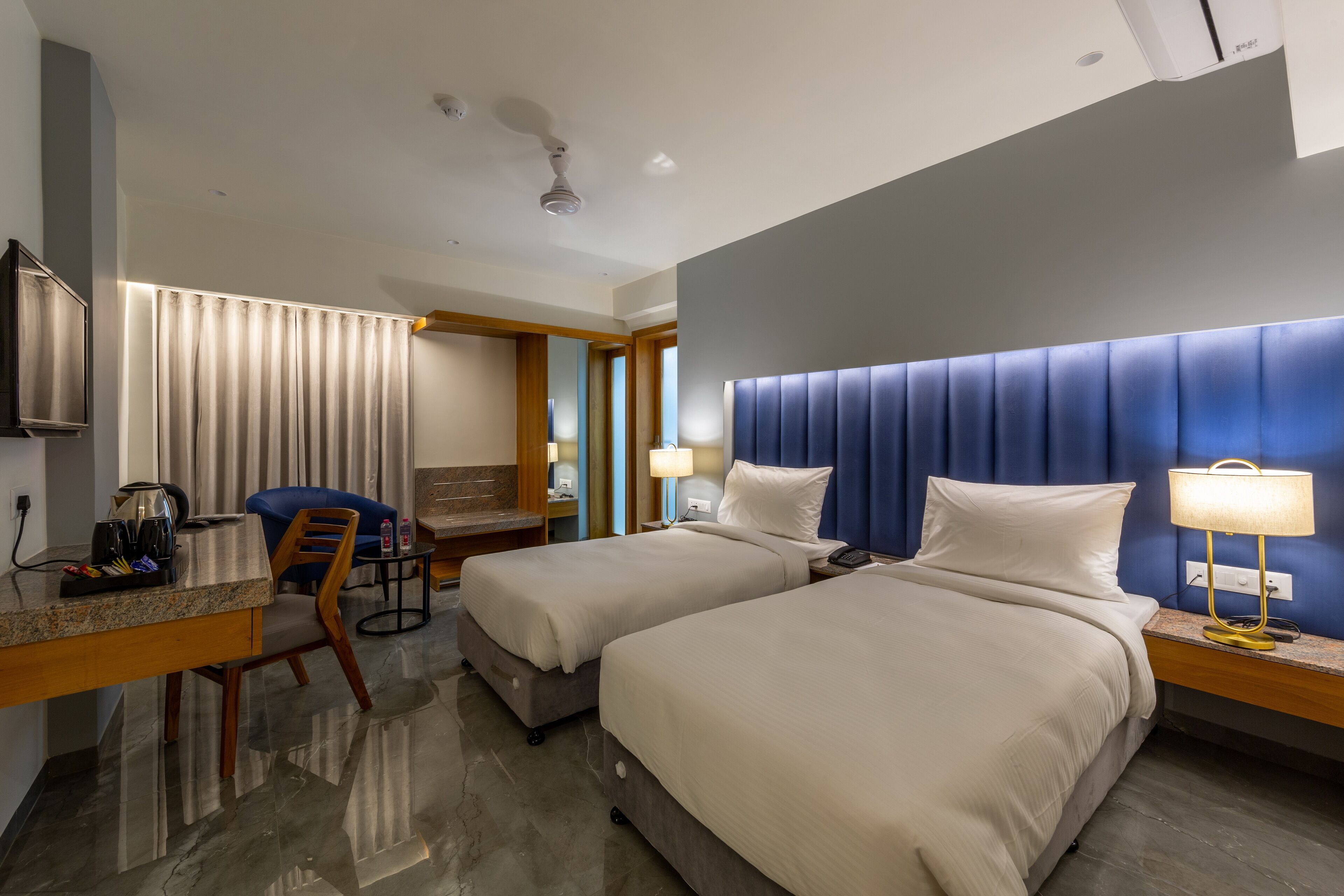 A hotel room with two beds, a desk, a chair, a TV, and a lamp.