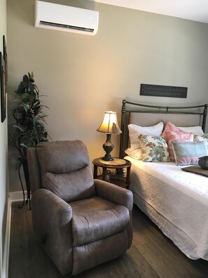 Un dormitorio con una butaca de cuero marrón, una mesita de noche con lámpara, una cama con ropa de cama floral y un aire acondicionado instalado en la pared.