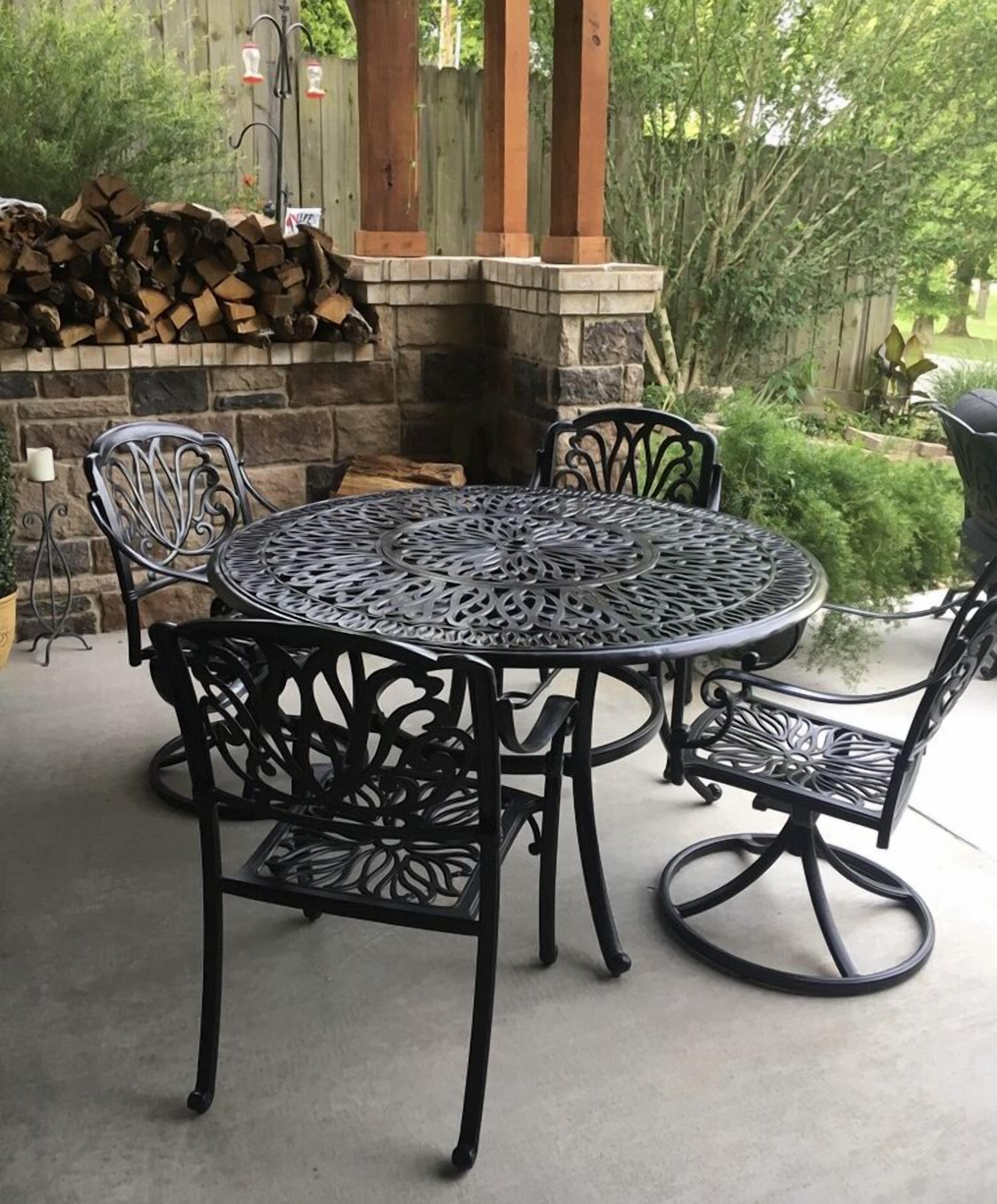 A round metal table with four chairs, set on a patio with a stone wall and a wooden fence in the background.