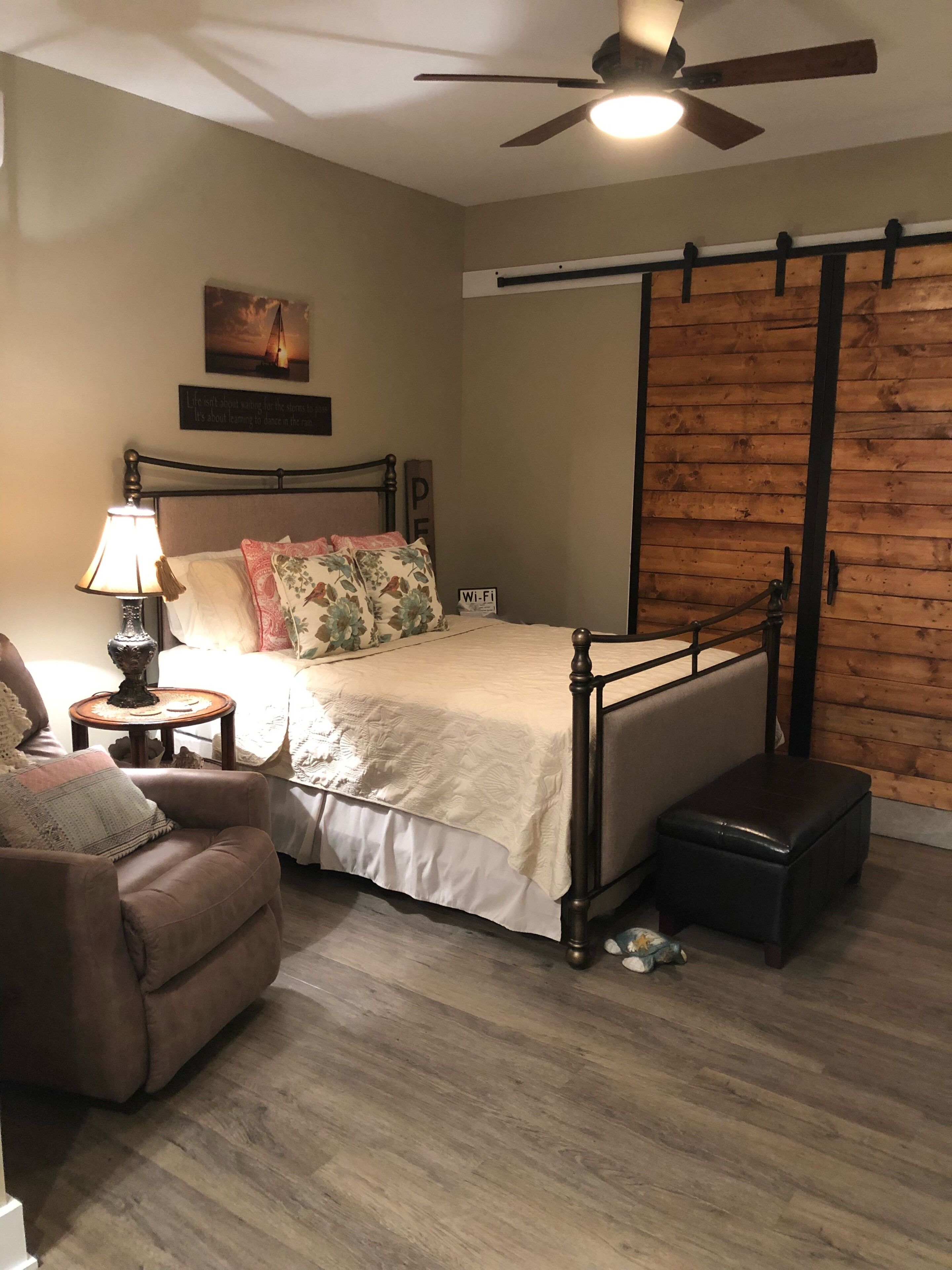 A bedroom with a bed, a chair, a nightstand, a lamp, a ceiling fan, and a wooden wall panel.