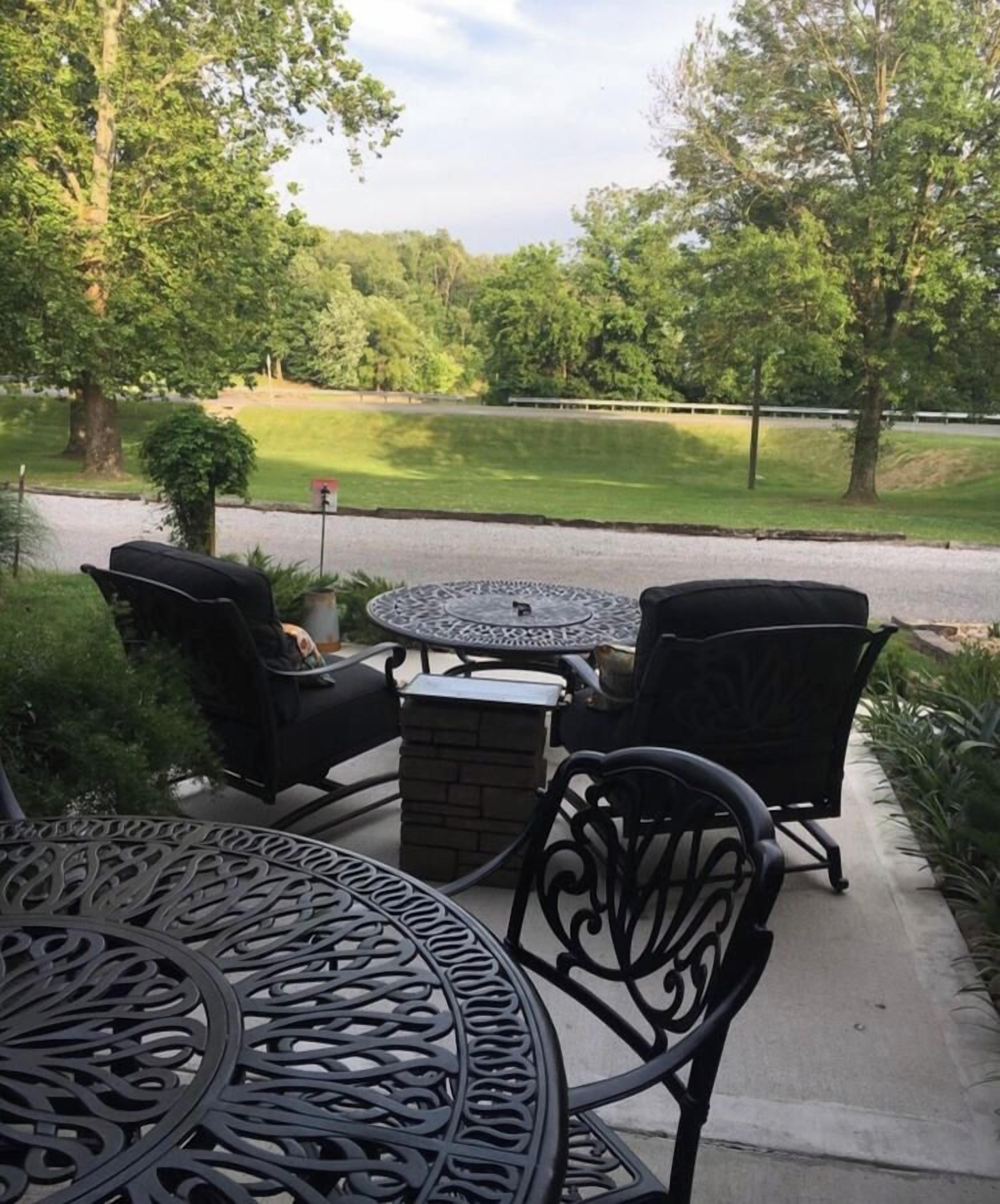 An outdoor patio with a round table and four chairs, surrounded by greenery and a view of a road.