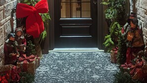 A stone-walled entrance with a dark wooden door, decorated with greenery and lights, leading to a carpeted area with Christmas decorations.