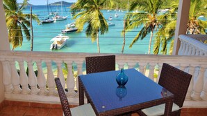 Un balcon avec une table et des chaises, donnant sur une marina où l’on peut admirer des bateaux et des palmiers.