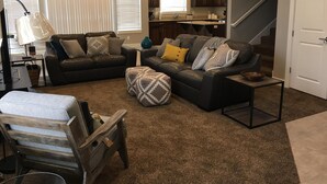 Ein Wohnzimmer mit Ecksofa, Hocker und einem Holzstuhl. Durch einen offenen Durchgang ist eine Küche mit Arbeitsfläche und Schränken zu sehen.