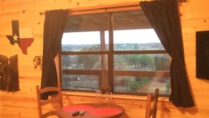 A wooden cabin interior with a large window, a table with chairs, and a Texas flag on the wall. - New lower rates. Great reviews   Elevated deck, Pet friendly, Romantic scenic (Fredericksburg)