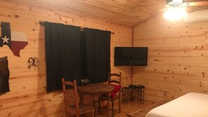 A cozy room with wooden walls, a ceiling fan, a TV mounted on the wall, a wooden table with chairs, and a bed with a white cover. - New lower rates. Great reviews   Elevated deck, Pet friendly, Romantic scenic (Fredericksburg)