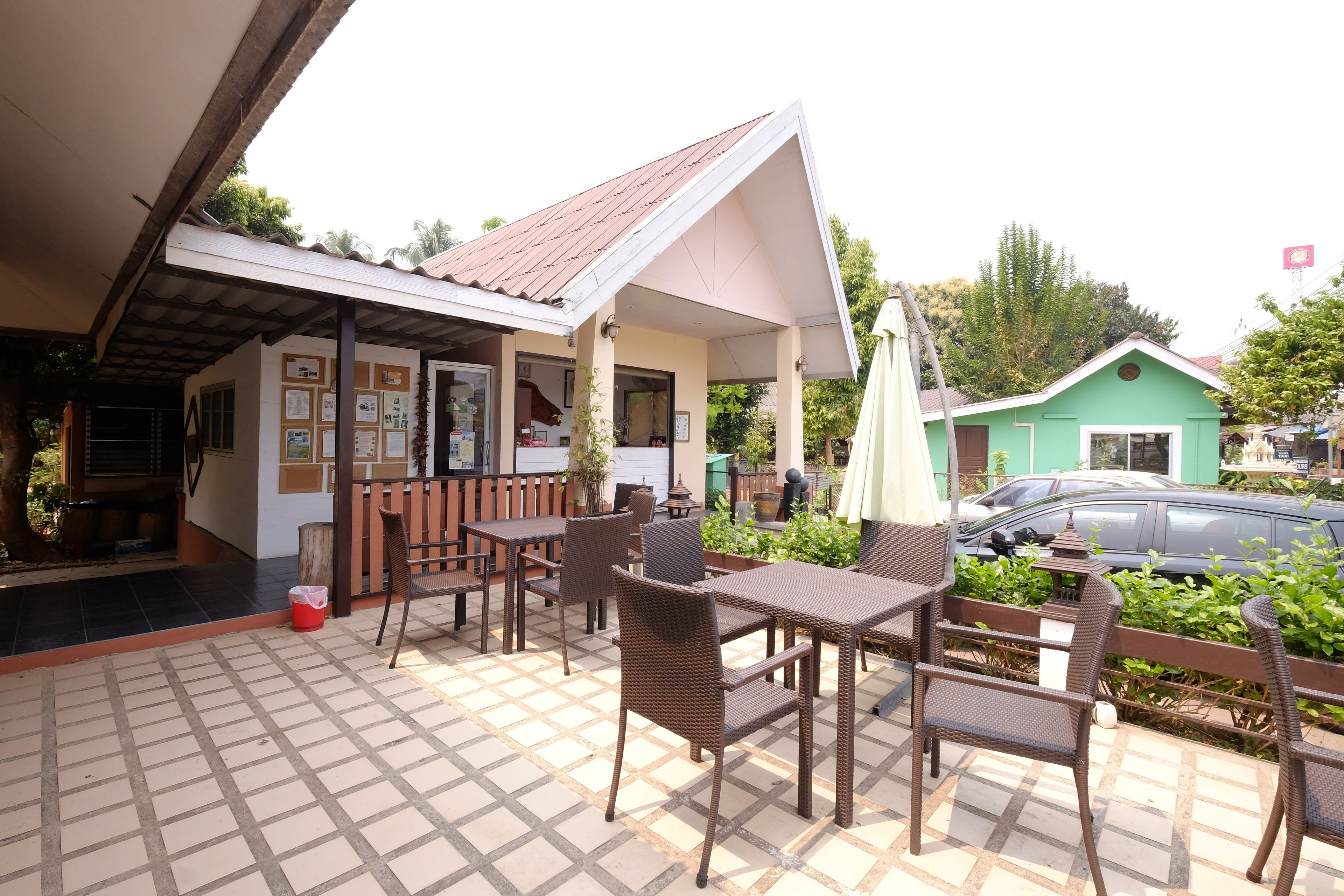 A covered outdoor seating area with tables and chairs, a small building with a tiled roof, and a green house in the background.