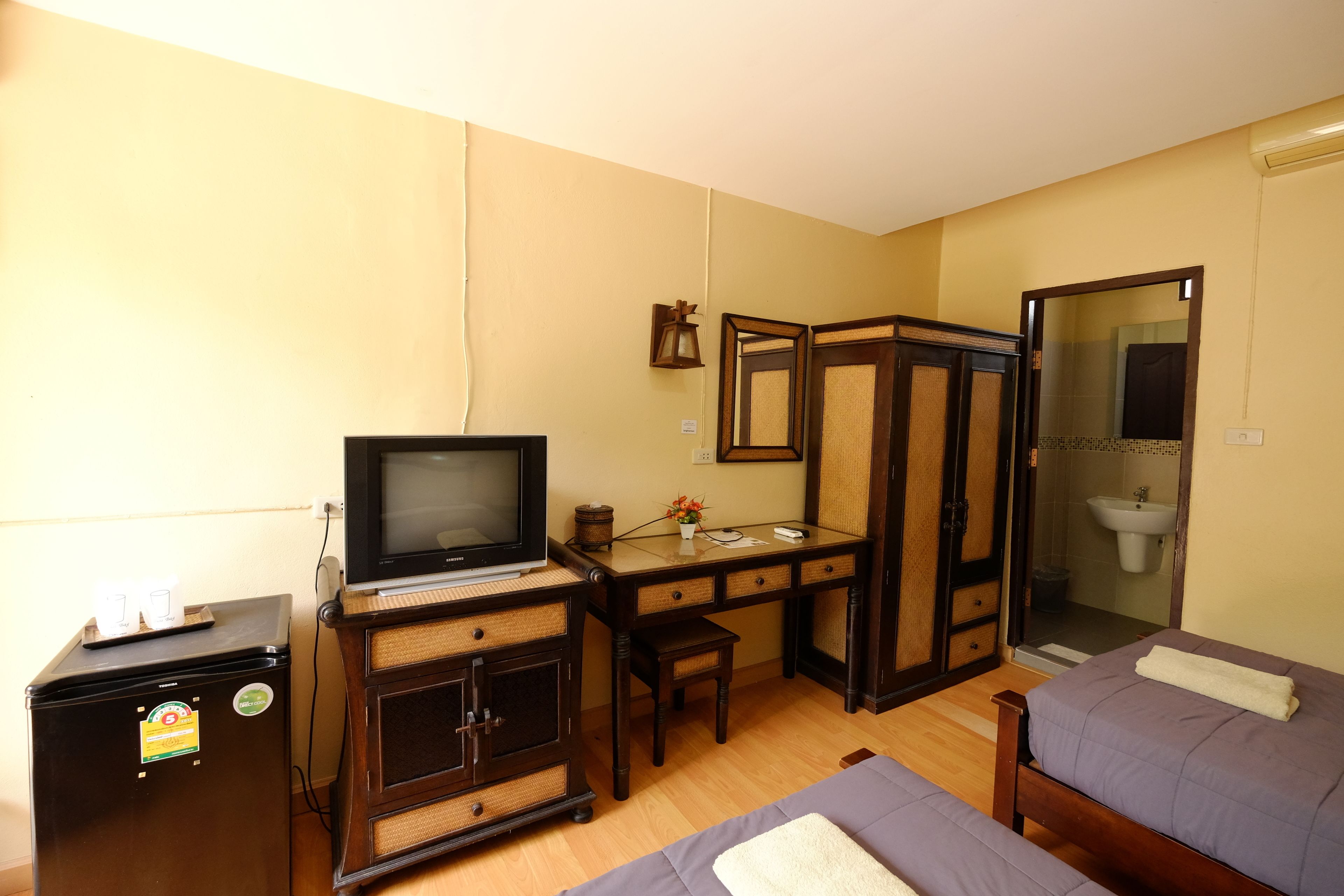 Une chambre d’hôtel de taille modeste, comprenant un lit simple, un bureau avec un ordinateur, une télévision et un petit réfrigérateur.