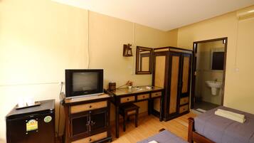 Une chambre d’hôtel de taille modeste, comprenant un lit simple, un bureau avec un ordinateur, une télévision et un petit réfrigérateur.