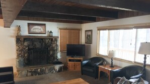 A living room with a stone fireplace, a television, a leather sofa, a wooden coffee table, and a window with blinds. - Walking distance to Snow Summit Resort. (Big Bear Lake)