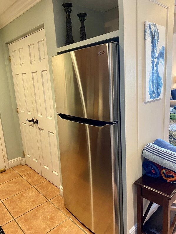 A stainless steel refrigerator in a kitchen with tiled flooring, a white door, and a small wooden table with a towel.