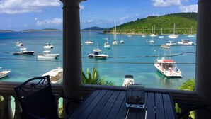 A view from a covered area with a table and chairs, overlooking a marina with various boats and a hilly landscape in the background. - Grande Bay Resort - Mojo Blue Luxury Studio Beachfront! (Cruz Bay)