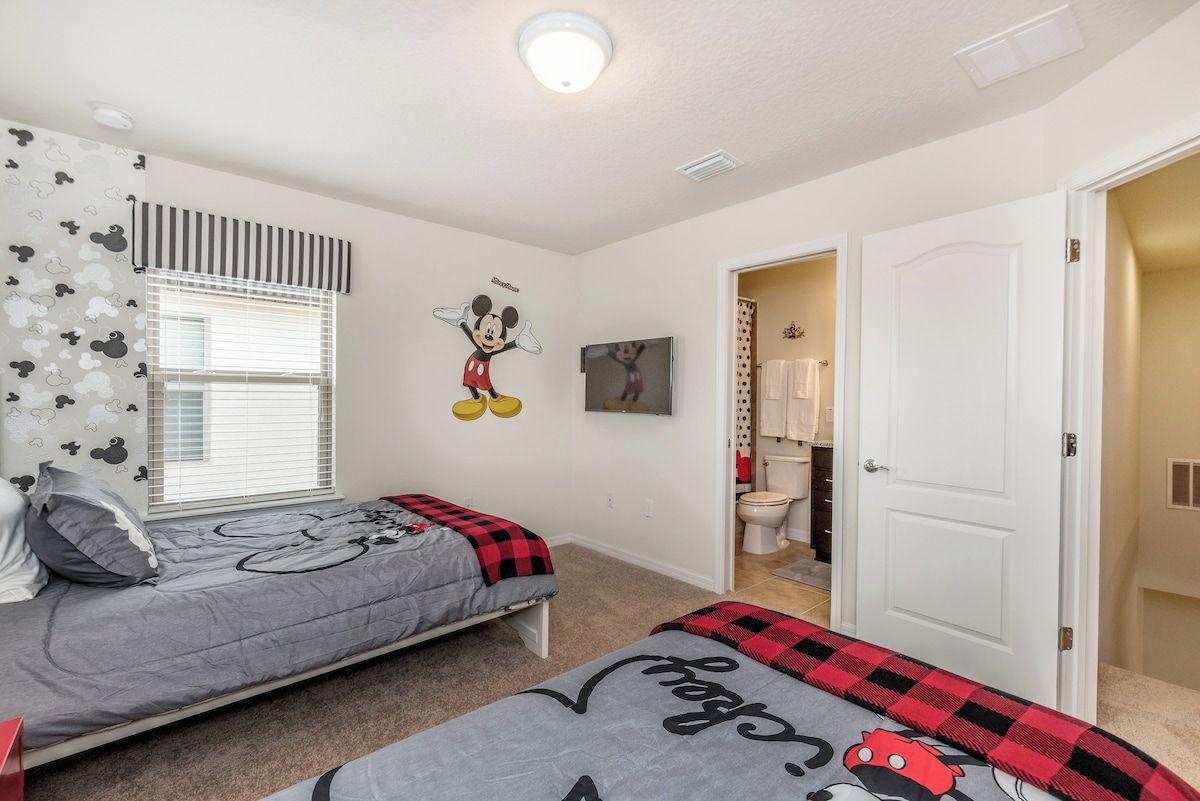 Una camera da letto con due letti, un adesivo a muro raffigurante Topolino, una persiana a righe e una porta bianca che dà su un bagno.