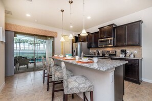 A modern kitchen with dark cabinetry, a central island with bar stools, and a dining area with a view. - Vacation Home: 6BR w/ Pool + Game Room near Disney (Kissimmee)