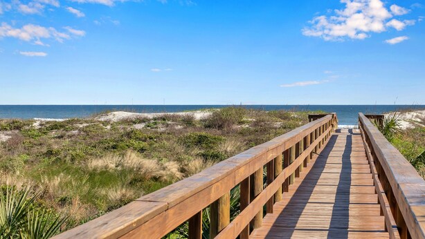 A wooden boardwalk leading to a beach with sand dunes and sparse vegetation.