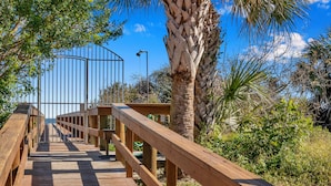 A wooden boardwalk leading to a gated entrance surrounded by palm trees and greenery.