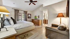 Une chambre à coucher comprenant un lit, une table de chevet, une lampe, une commode, un téléviseur et donnant sur la cuisine.