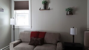 Ein Wohnzimmer mit einem beigen Sofa, einem roten Sitzkissen, einer Stehlampe und zwei an der Wand befestigten Regalen mit Pflanzen.