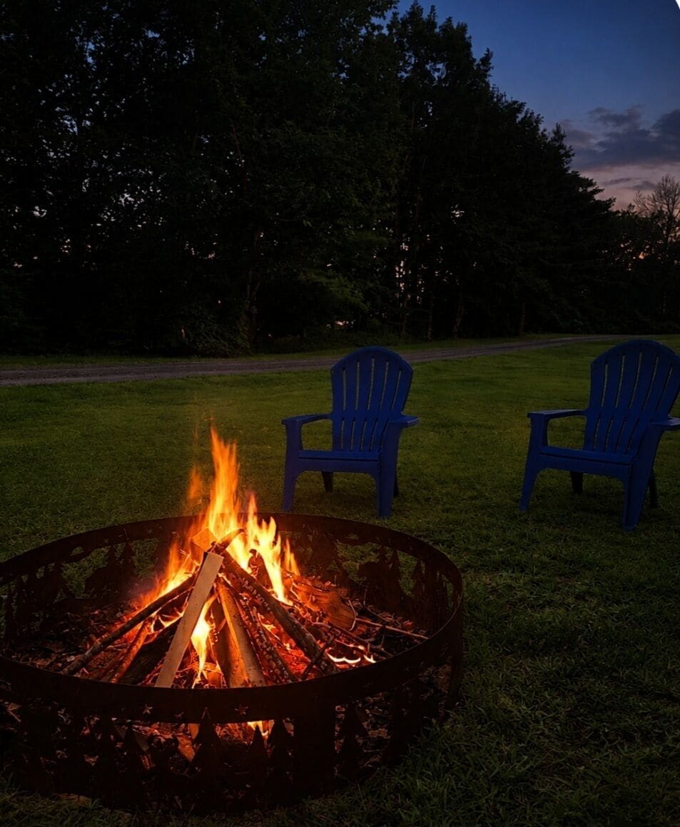 Ein Lagerfeuer mit brennenden Holzscheiten, umgeben von Gras, zwei blaue Stühle und eine Straße im Hintergrund.