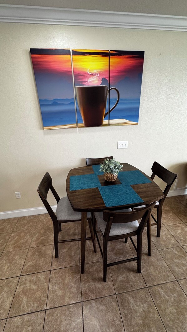 A dining area with a wooden table and four chairs, a blue placemat, and a potted plant. A large painting of a sunset and a cup hangs on the wall above the table.
