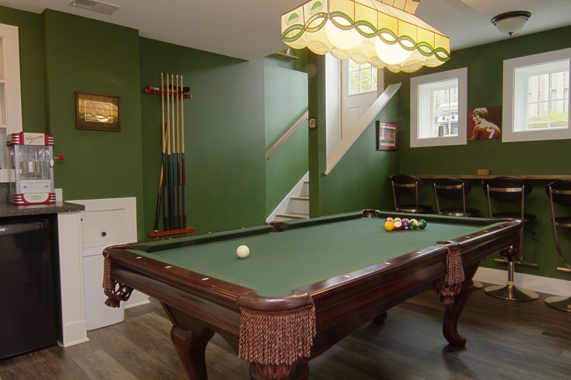 A pool table with a green felt surface, surrounded by a green wall, a staircase, and a bar area with chairs.