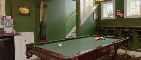 A pool table with a green felt surface, surrounded by a green wall, a staircase, and a bar area with chairs.