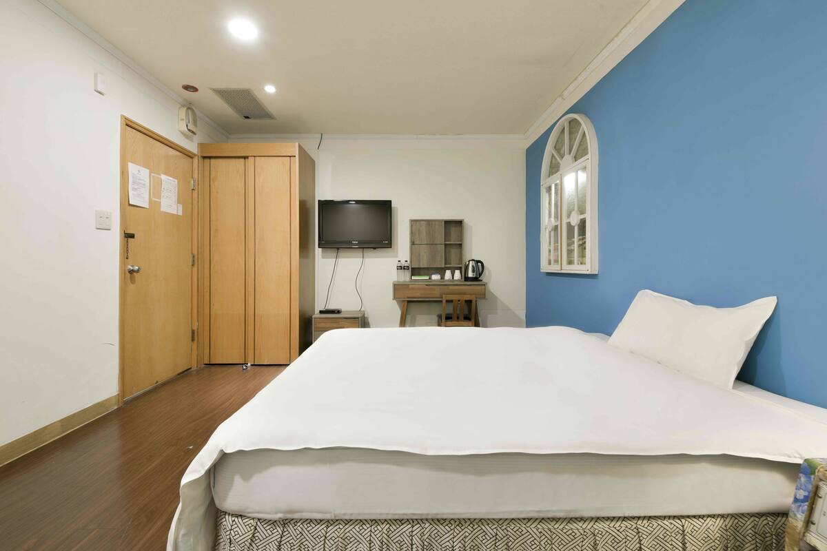 A hotel room with a large bed, a wooden wardrobe, a television, a desk with a chair, and a window with a view.