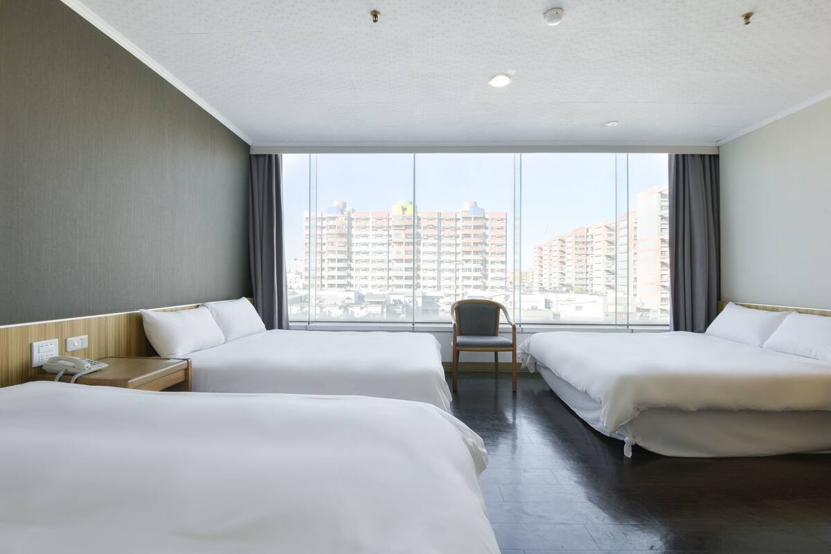 A hotel room with two beds, a chair, a bedside table with a phone, and a large window offering a city view.