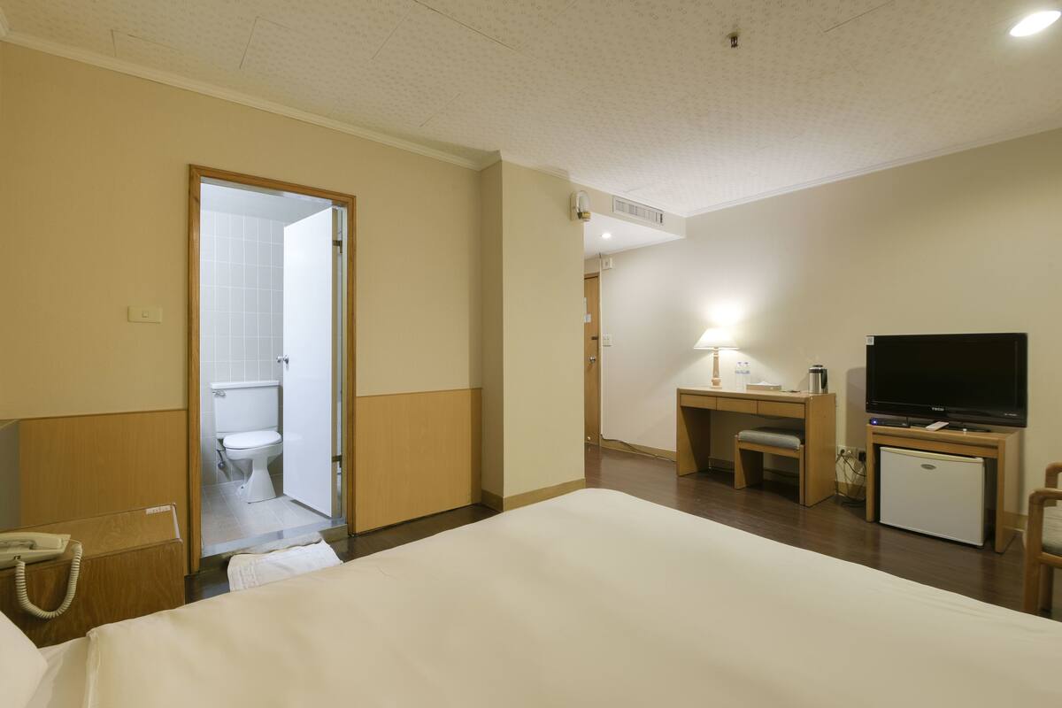 A hotel room with a bed, a desk with a chair, a television, a mini-fridge, and a bathroom visible through an open door.