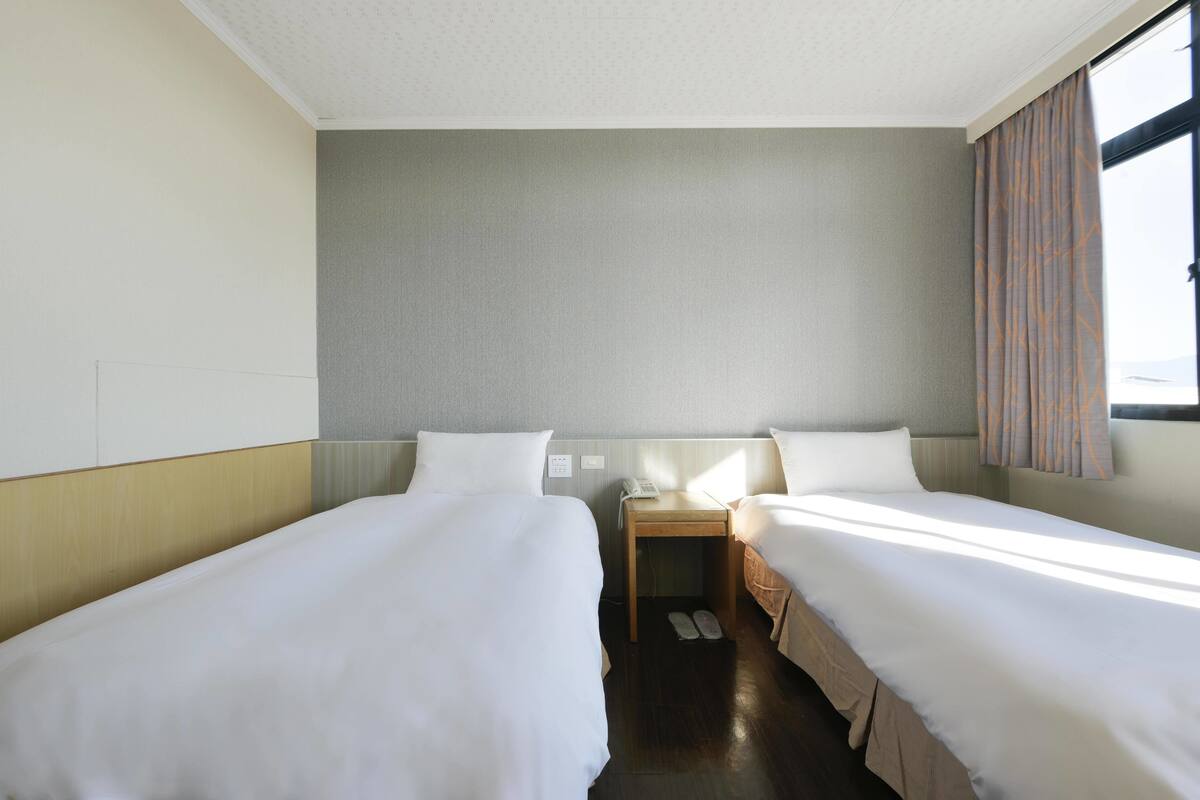 A hotel room with two beds, a wooden nightstand, and a window with curtains.