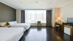 A hotel room with two beds, a desk, a chair, a TV, and a view of buildings through the window. - Back Home Hotel (Chiayi City)