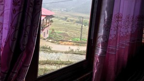 A window with purple curtains, a view of a rural landscape with terraced fields, a building with a red roof, and distant hills. - Moc Anh House (Ta Van)