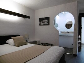 Quarto com cama, mesa de cabeceira, abajur, quadro na parede e banheiro visível por um arco.