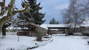 A snow-covered landscape with a wooden building, a paved driveway, and a gate. - Relais Des Vieilles Postes (Les Avenieres-Veyrins-Thuellin)