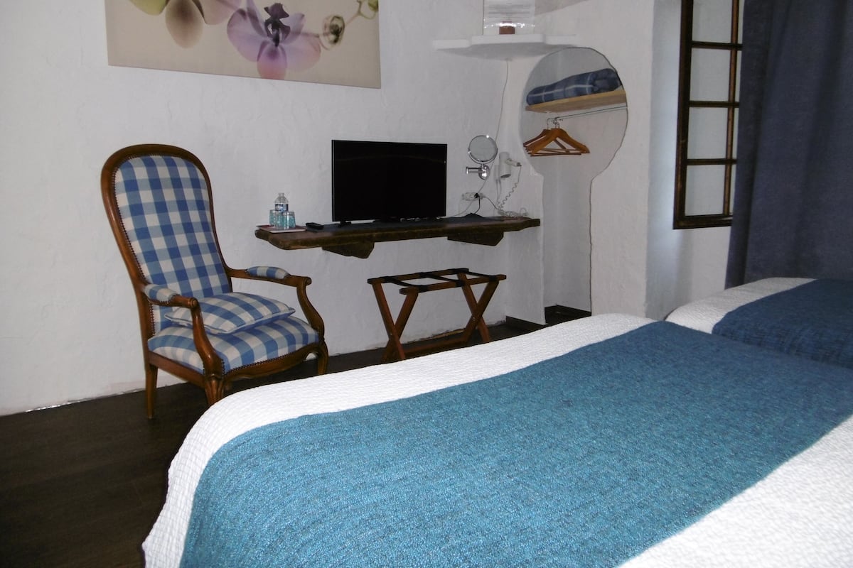 Une chambre à coucher avec un lit, une chaise, un bureau avec un ordinateur, un miroir et une fenêtre avec des rideaux.