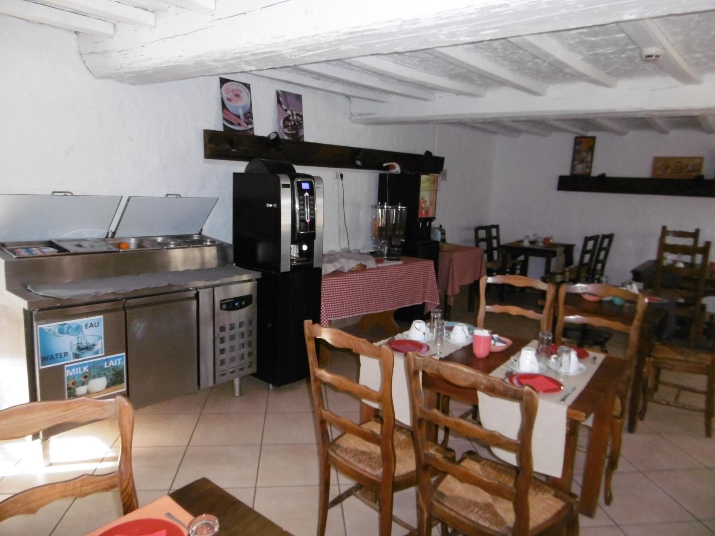 On aperçoit un coin repas avec des chaises en bois et une table dressée pour deux personnes. Au fond, on distingue également un coin cuisine avec un réfrigérateur et un distributeur d’eau.