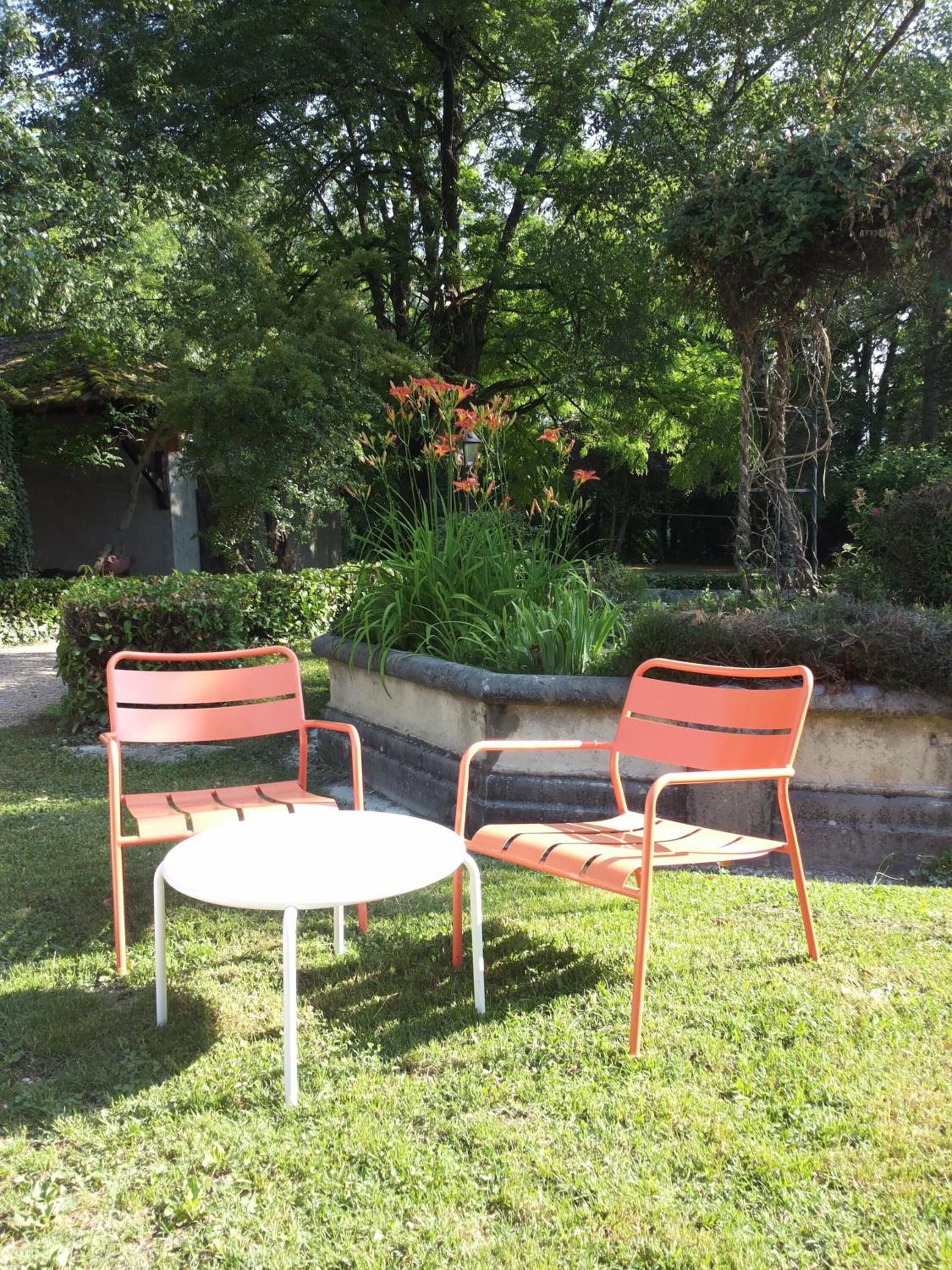 An outdoor seating area with two metal chairs and a small round table, surrounded by lush greenery and trees.