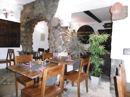 Interior de restaurante com paredes de pedra, mesas e cadeiras de madeira e uma grande planta em vaso.