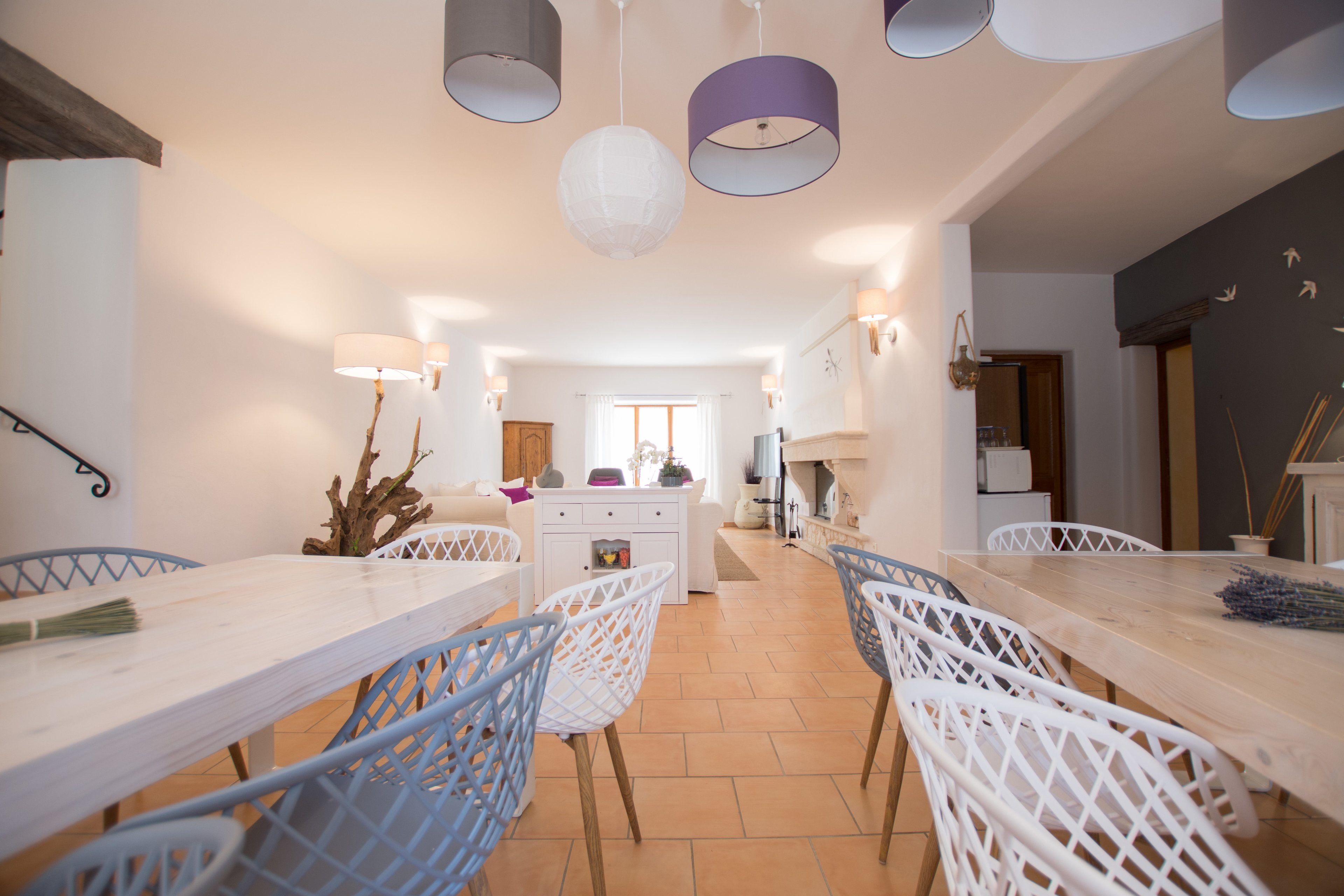 Un comedor amplio con una larga mesa y sillas blancas, una cocina al fondo, e iluminación colgante.